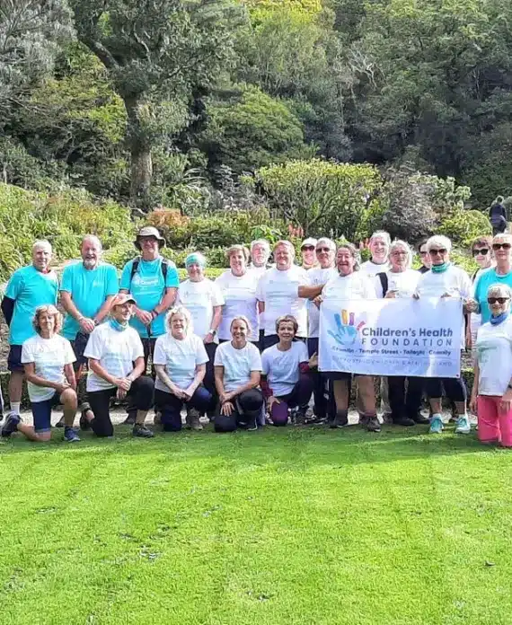 A group of adults pose outdoors in a garden, standing and kneeling on grass. Many wear white or turquoise T-shirts. Several hold a "Children's Health Foundation" banner. Trees and greenery are in the background.
