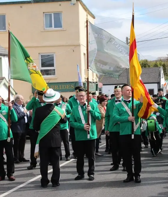 A marching band in green jackets and hats performs in a parade, carrying colourful flags. Spectators line the street, watching the lively event in a small town with buildings in the background.