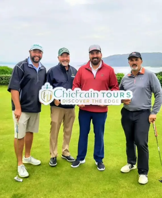Four men stand on a golf course holding a "Chieftain Tours Discover the Edge" sign. Three hold golf clubs, and they are all smiling at the camera with green hills and water visible in the background.