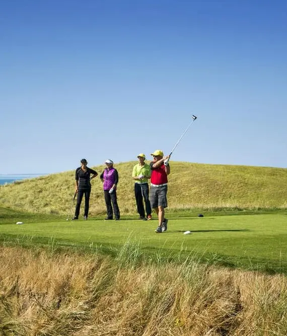 A group of people stand on a golf course under a clear blue sky; one person in a red shirt swings a golf club whilst others watch, surrounded by grassy hills and distant water.