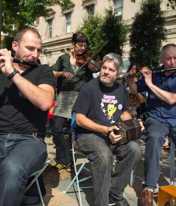 Three musicians perform outdoors; two play flutes whilst seated, and one plays a concertina. A violinist stands behind them. People watch and children stand nearby on a sunny day in an urban setting.