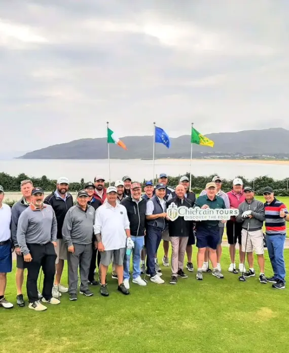 A group of men pose together on a golf course, some holding golf clubs and a "Chefcrain Tours" sign. Three flags and a scenic landscape with hills, water, and cloudy skies are visible in the background.