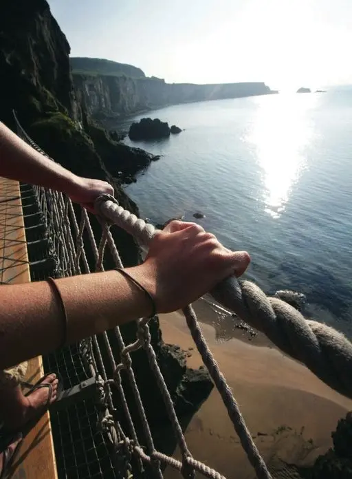 A person holds onto a thick rope bridge handrail above a rocky coastline, overlooking calm sea waters and a distant cliff under a clear, sunny sky.