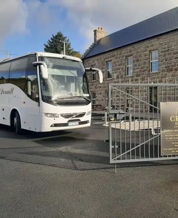 A white tour coach is parked outside the entrance gate of Crolly Distillery, which is housed in a stone building. A sign on the gate displays the distillery’s name and visitor information.