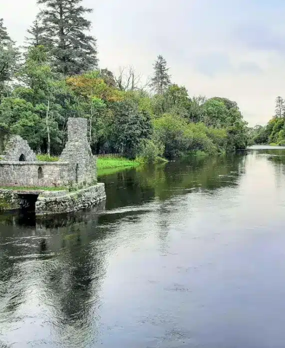 A small stone ruin stands by the edge of a calm river, surrounded by dense green trees and grass under a cloudy sky. The water reflects the trees and the remains of the structure.