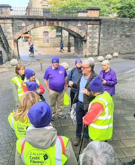 A group of people wearing high-visibility vests and purple hats, some with "Cystic Fibrosis Trust" logos, stand in a circle outside near a stone archway, listening to a man speak.
