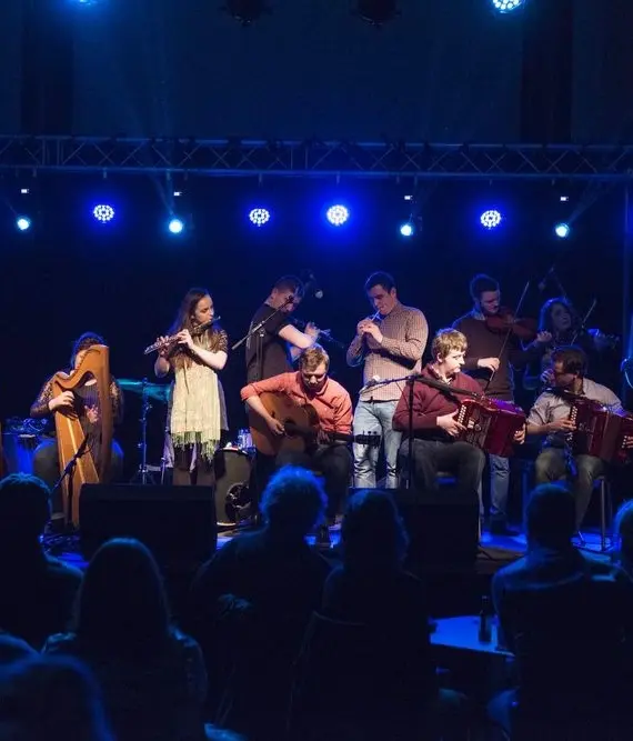 A group of musicians perform on a stage under blue lights, playing various instruments such as harps, flutes, guitars, and accordions, whilst an audience watches from the foreground.