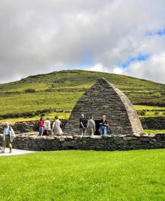 Tourists visit the ancient stone Gallarus Oratory, surrounded by lush green grass and hills under a partly cloudy sky in Ireland. Some people are walking, whilst others stand near the structure.