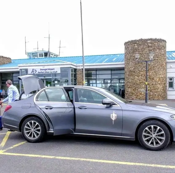 A silver saloon with its driver's door open is parked outside an airport terminal. Three people with suitcases stand nearby, and the terminal building is visible in the background.
