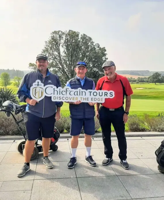 Three men in golf attire stand on a golf course patio, smiling and holding a "Chieftain Tours Discover The Edge" sign. Two golf bags on trolleys are beside them, with green fairways and trees in the background.