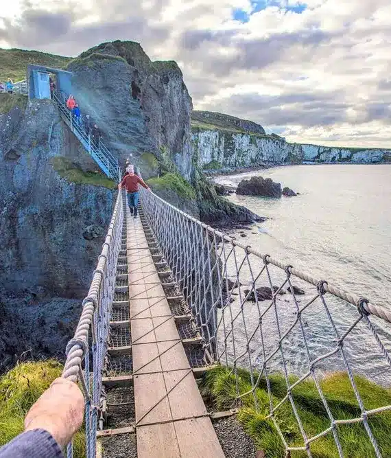 A person crosses a narrow rope bridge suspended over cliffs and the sea, leading to a grassy rocky island under a partly cloudy sky. Other people are ahead on the path.