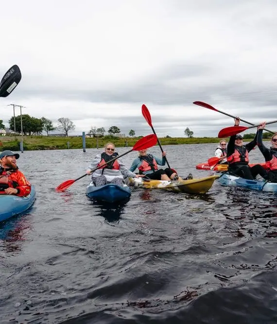 A group of people kayaking together on a calm body of water, all wearing life jackets and holding paddles up, smiling and appearing cheerful under a cloudy sky near a shoreline with houses.