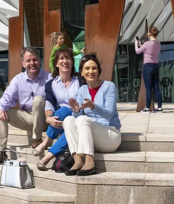 Three people sit smiling on outdoor steps, posing for a photo on a sunny day; two women and one man are in the foreground, with other people and a modern building in the background.