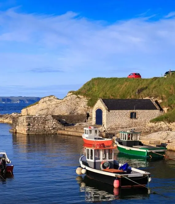 Small boats are moored in a calm harbour surrounded by stone walls and grassy hills, with a stone building and a red car on the road above, under a bright blue sky with scattered clouds.