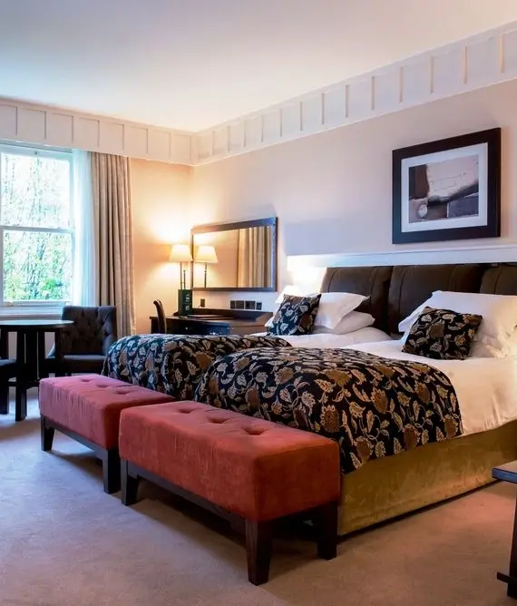 A spacious hotel room with a large double bed, patterned bedding, two red pouffes, dark wood furniture, a table with chairs by the window, lamps on bedside tables, and framed art on the wall above the bed.