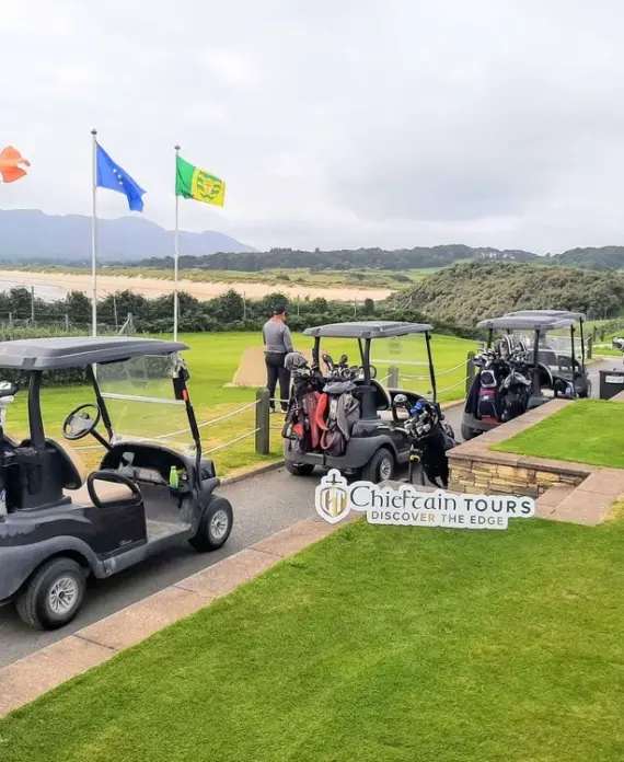 Three golf buggies with golf bags parked on a grassy area beside a golf course; three people stand nearby. Irish, European Union, and “SF” flags fly in the background. A Chieftain Tours sign is visible.
