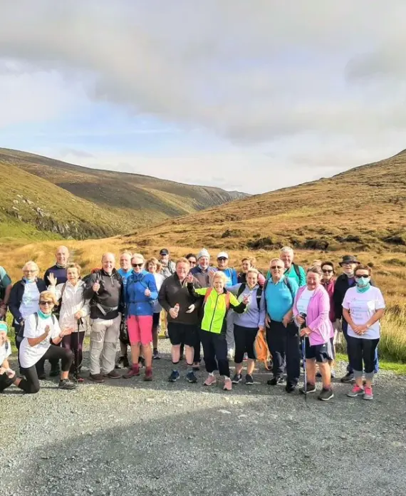 A group of people pose together on a gravel path in a hilly, grassy landscape under a cloudy sky, dressed in casual outdoor clothing and ready for a walk.