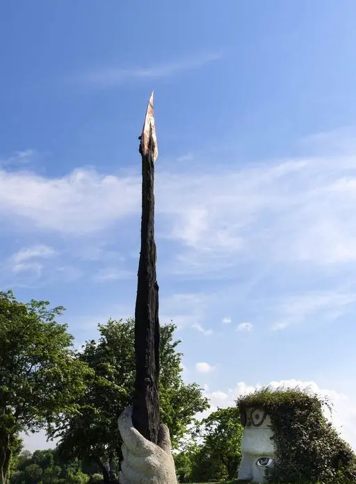 A tall, charred wooden sculpture with a pointed top rises from a white, hand-like base on a grassy field; nearby, a quirky bush is trimmed into a face with large eyes and a wide mouth. Trees and blue sky fill the background.
