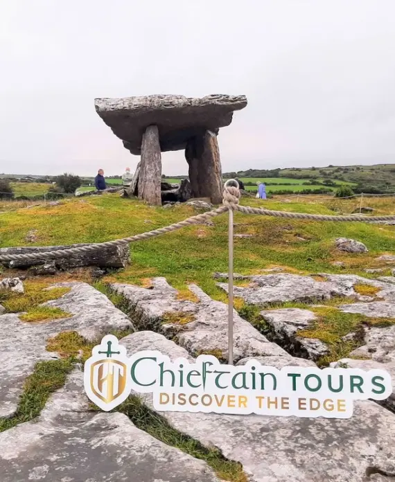A large ancient stone portal tomb stands on grassy, rocky ground under a cloudy sky. A rope barrier surrounds the site. "Chieftain Tours Discover the Edge" logo is visible at the bottom of the image.