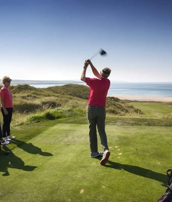 Three people in red shirts on a golf course by the sea. One person is swinging a golf club whilst the other two watch. Golf bags and grassy dunes surround the green, with blue sky and sea in the background.