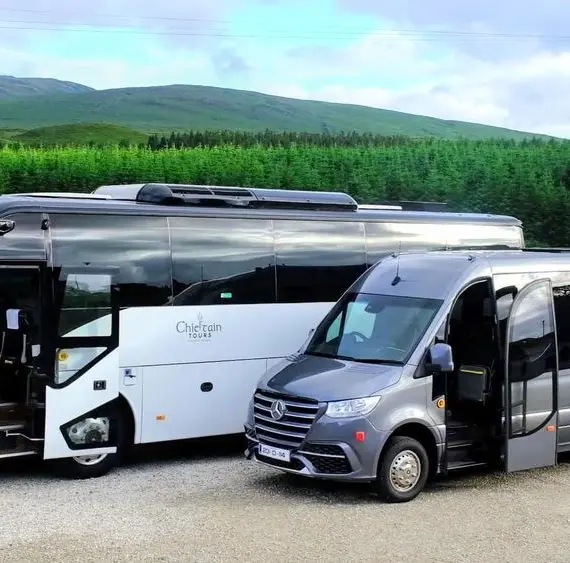 Two modern tour coaches, one large and one mid-sized, are parked side by side on a gravel car park with green hills, trees, and an Irish flag in the background. Both vehicles display "Chieftain Tours" on the side.