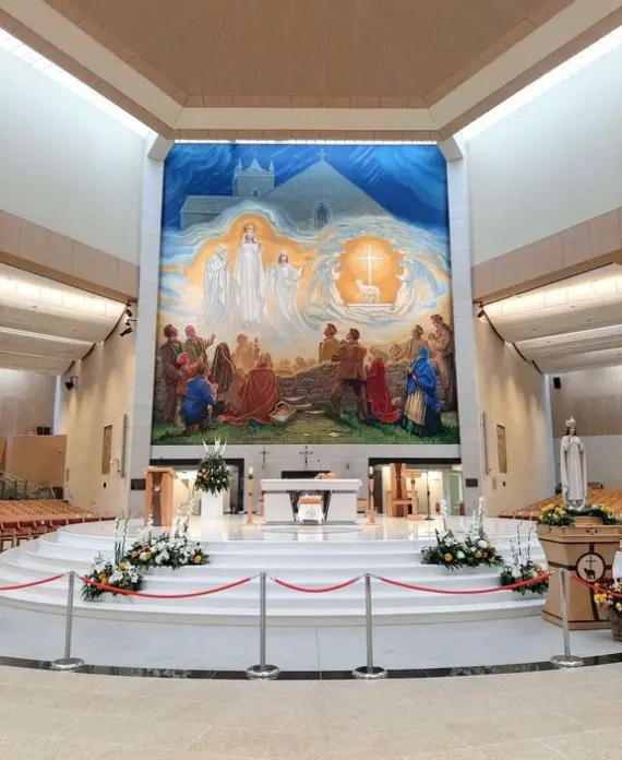 The interior of a modern church with a large, colourful mural behind the altar depicting a religious scene. The altar is decorated with white flowers, and rows of seating line both sides of the spacious sanctuary.