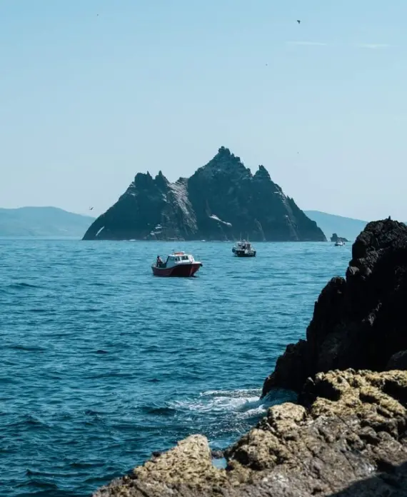 Small boats float on blue ocean water near a steep, rocky island under a clear sky. Jagged rocks are visible in the foreground, with distant mountains faintly on the horizon.