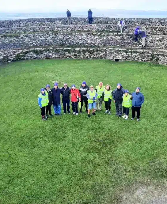 A group of people stands in a circle on green grass inside a large, ancient stone ring fort, with a few more people and layered stone walls in the background. The sky appears overcast.