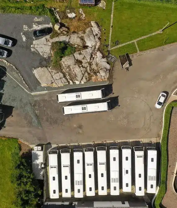 Aerial view of a car park with several white buses and vans parked in rows near a building with paved footpaths and green grassy areas.