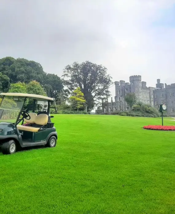 A golf buggy sits on a lush green lawn with a large stone castle and tall trees in the background under a partly cloudy sky.