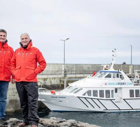Two men wearing red jackets stand on rocks near a moored white ferry with "DoolinFerry.com" written on it. The background shows a concrete wall and a cloudy sky.