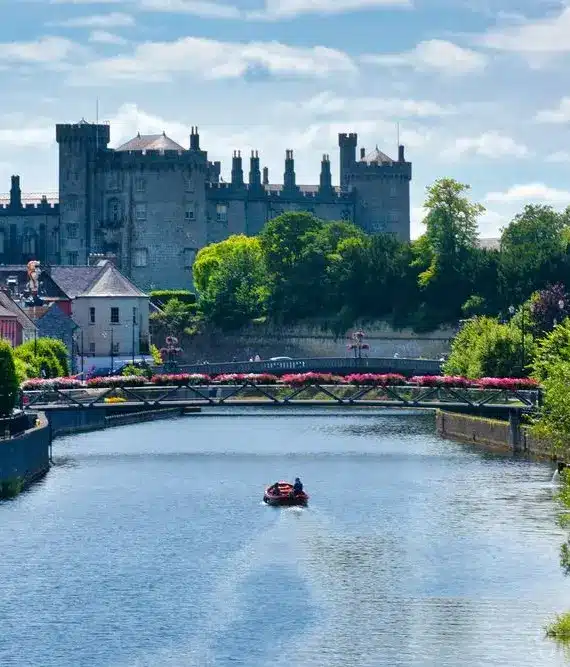 A river flows through a green, leafy town with a small boat in the water. In the background, a large, historic castle stands atop a hill. A bridge adorned with flowers crosses the river.