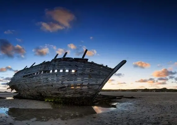 An old, weathered shipwreck rests on a sandy, deserted shore at sunset, with dramatic clouds and a blue sky in the background. Shallow water reflects part of the ship’s hull.