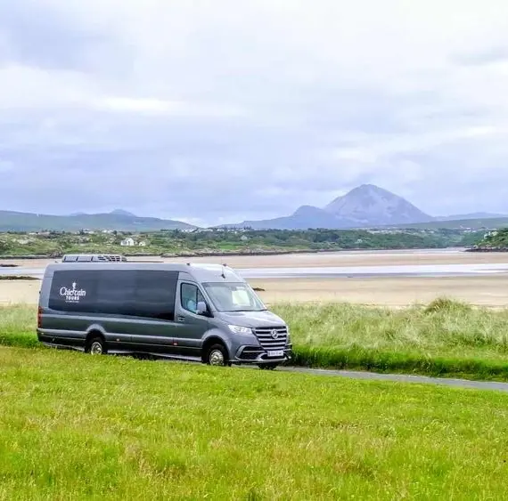 A dark grey camper van drives along a grassy road near a sandy beach, with green hills and a mountain in the background under a cloudy sky.
