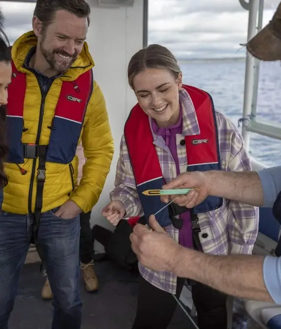 A group of people wearing life jackets and outdoor clothing smile and watch as a man holds up a small sea creature on a boat, with water visible through the windows.