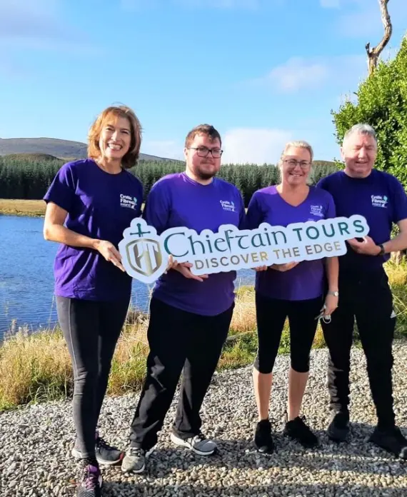 Four people in matching purple shirts stand outdoors by a lake, holding a "Chieftain Tours Discover the Edge" sign. There are trees, grass, and hills in the background under a blue sky.