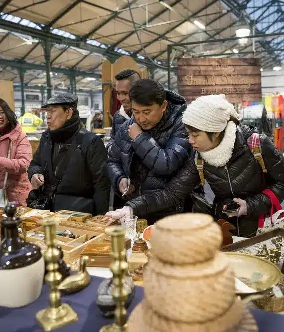 A group of people wearing winter jackets browse various items, including baskets, vases, and wooden objects, at an indoor market with a high metal roof. Some are closely examining the merchandise on display.