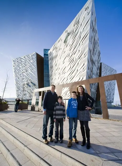 A family of four stands smiling in front of the modern, angular Titanic Belfast museum building on a sunny day with a clear blue sky.