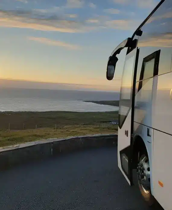 A tour bus with "Chieftain Tours" written on the side is parked near a coastal road, overlooking the sea at sunset with grassy land stretching towards the water.