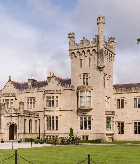 A large stone castle with multiple towers and arched windows, surrounded by a manicured lawn and trees, under a partly cloudy sky.