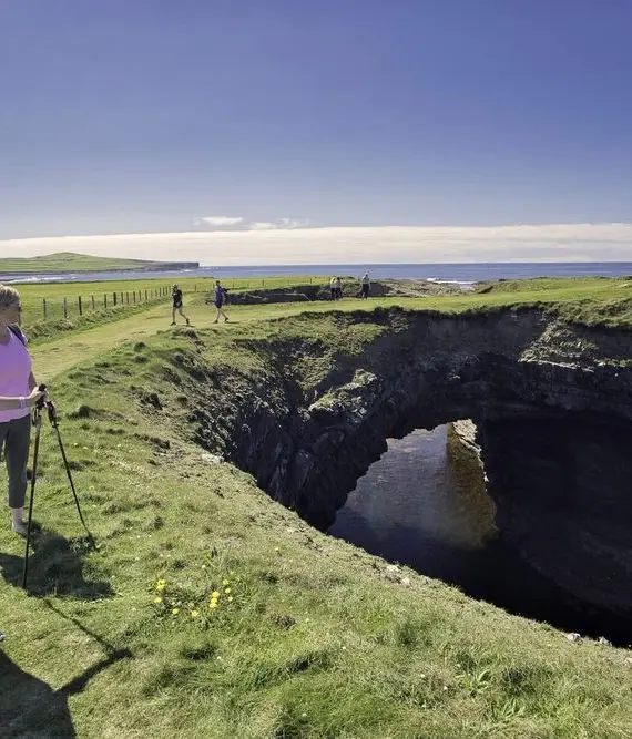Three people stand on grassy cliffs near a large coastal hole or cave, looking at the view under a clear blue sky; other walkers are visible in the background.