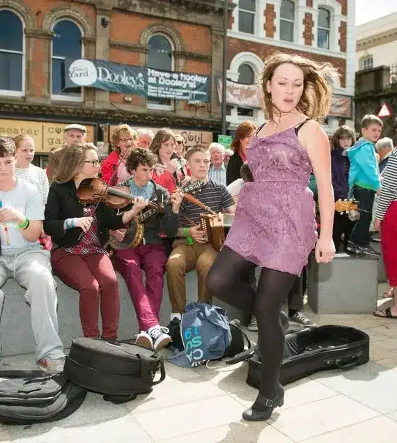 A lively street scene shows musicians playing traditional instruments as a woman in a purple dress and a young girl dance. A crowd watches, some smiling, on a sunny day in front of brick and white buildings.