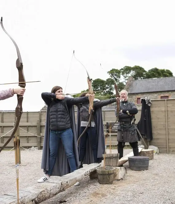 Three people stand outdoors in front of a wooden fence, drawing bows and aiming arrows. Two wear cloaks, resembling medieval costumes, and one wears modern clothing. A stone building is visible in the background.
