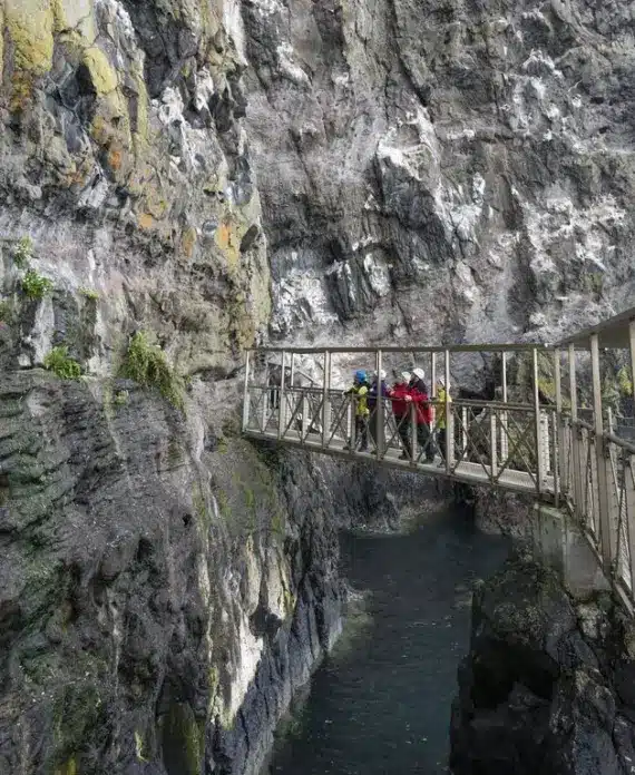 People wearing helmets and outdoor gear walk across a narrow metal bridge attached to a steep rocky cliff above a body of water in a gorge.