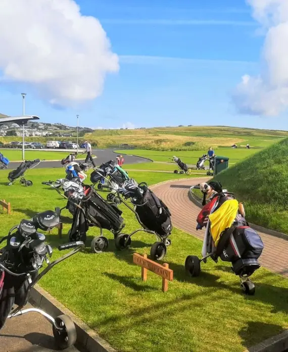 Several golf trolleys with golf bags are parked on grass near a pavement at a golf course, with golfers preparing in the background under a partly cloudy sky.