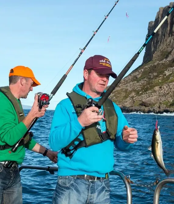 Two men fishing from a boat; one in a green jacket and orange cap holds a fishing rod, while the other in a blue jacket and life vest has caught a fish. Rocky coastline and blue water are in the background.