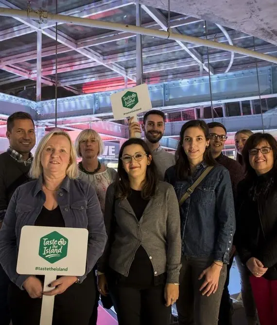 A group of people pose together indoors, smiling at the camera. Two people in front hold signs that read "Taste the Island." The background features modern industrial architecture with glass and metal elements.