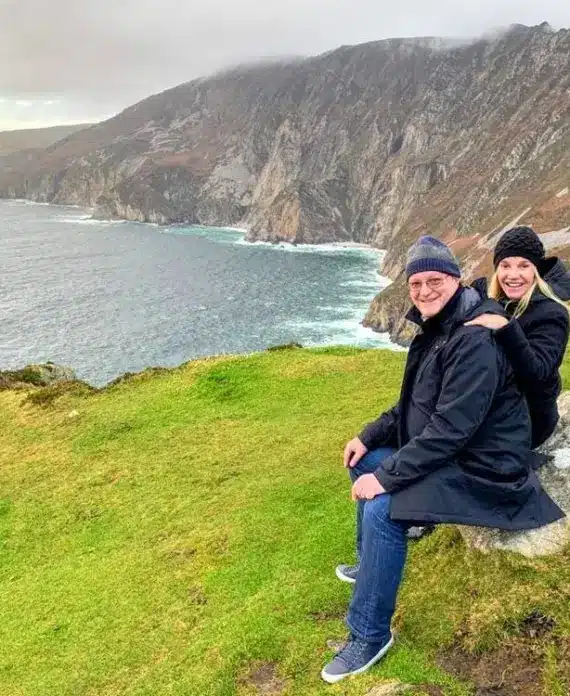 A smiling couple in black jackets and hats sit on a grassy cliff overlooking dramatic sea cliffs and the sea, with cloudy skies and rugged mountains in the background.