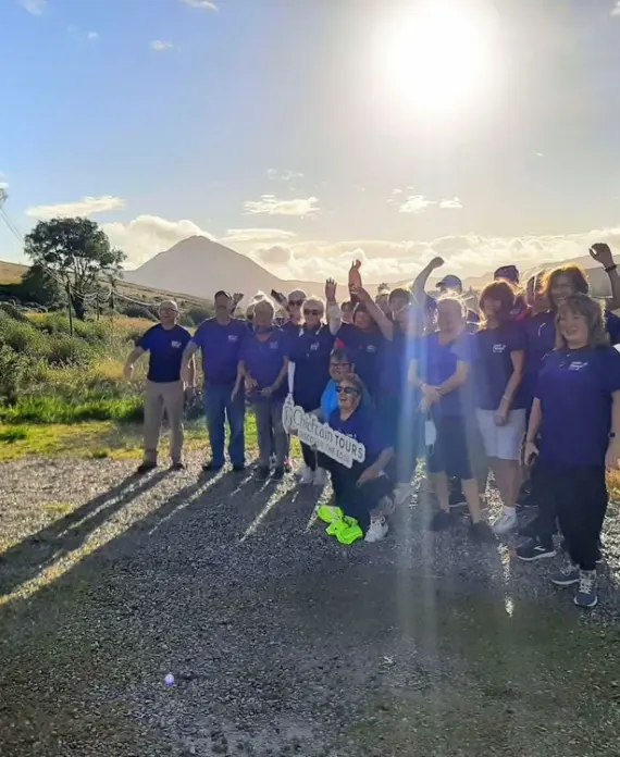 A group of people in matching blue shirts pose together outdoors by a lake with the sun shining brightly behind them, casting long shadows. Some people hold a white sign and trees and hills are visible in the background.