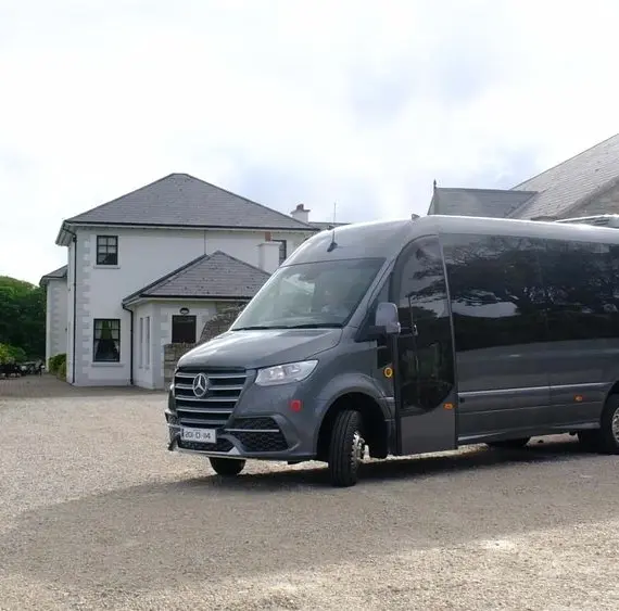 A dark grey Mercedes-Benz tour van labelled "Chieftain Tours" is parked on a gravel drive in front of a large, white house with grey roofing on a cloudy day.